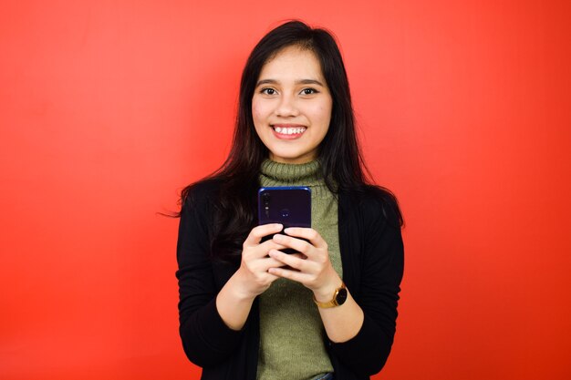 À l'aide d'un smartphone et souriant de jeunes belles femmes asiatiques utilisant un pull noir isolé sur rouge