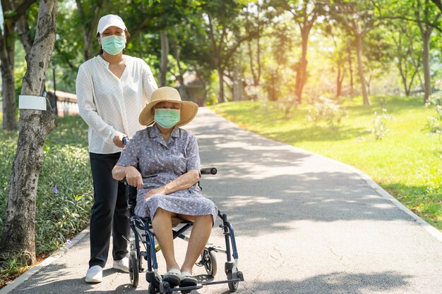 Aide et soins du soignant femme âgée asiatique ou âgée femme patiente assise sur un fauteuil roulant et portant un masque facial pour voyager dans le parc