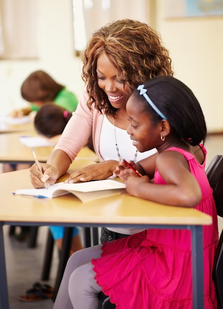 Aider l'enseignement et l'enseignant avec un enfant à l'école pour l'apprentissage ou le développement de l'éducation Heureuse femme noire parlant à un élève de l'écriture de cours et des connaissances à un bureau de classe avec soutien