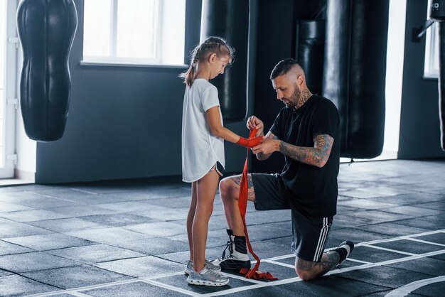 Aider à Porter Les Bandages. Le Jeune Entraîneur De Boxe Tatoué Enseigne La Petite Fille Mignonne Dans Le Gymnase.