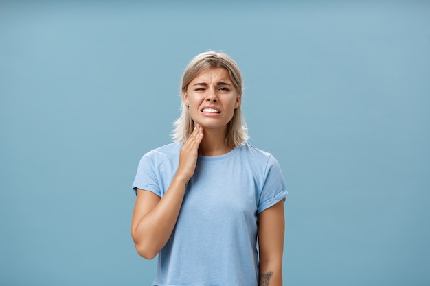 Aïe ma gorge. Mécontent malheureux belle fille blonde adulte avec une coupe de cheveux courte serrant les dents d'une sensation douloureuse touchant le cou ayant une angine ou une inflammation posant