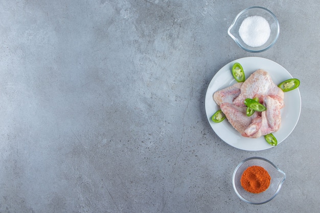 Ailes de poulet sur une assiette à côté de bols d'épices et de sel, sur le fond de marbre.