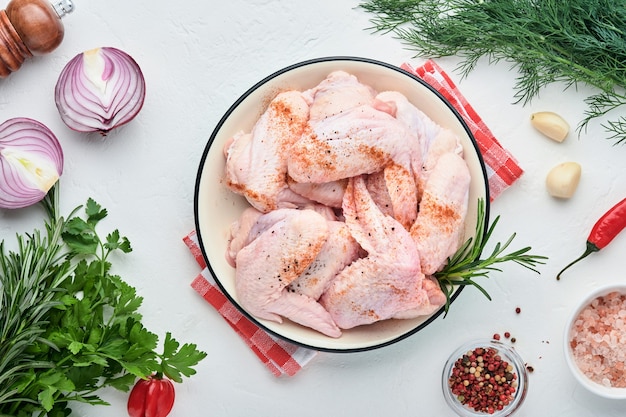 Ailes de poulet crues dans un bol blanc avec des épices et des ingrédients pour cuisiner sur fond d'ardoise claire, de pierre ou de béton. Vue de dessus avec espace de copie. Maquette.