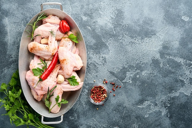 Ailes de poulet crues dans une casserole en métal ou un bol avec des épices et des ingrédients pour la cuisson sur une surface de béton gris foncé. Vue de dessus.