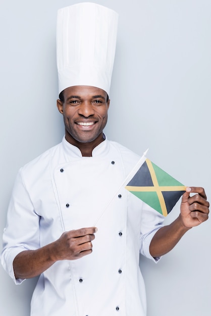 aimer cuisiner des plats nationaux. jeune chef africain confiant en uniforme blanc tenant le drapeau de la jamaïque
