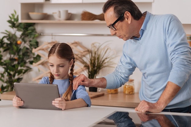 Aimer le grand-père vieillissant attentionné debout à la maison tout en exprimant ses soins et en retirant la tablette de sa petite-fille