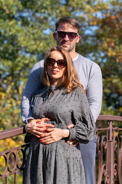 Aimer le jeune couple posant sur un pont en fer forgé à l'extérieur dans un parc d'automne debout dans une étreinte étroite souriant à la caméra