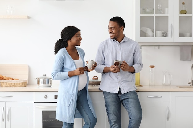 Aimer s'attendre à ce que la famille noire boive du café dans la cuisine, se parle et se regarde. Joyeuse femme enceinte afro-américaine et son mari avec des tasses, dégustant une tisane avec des biscuits