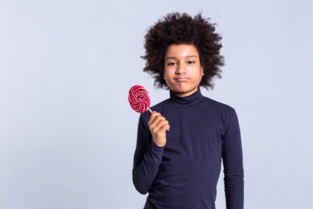 L'air extrêmement fier. Charmant jeune garçon à la peau foncée, sceptique tout en portant des bonbons sur un bâton en bois dans une main