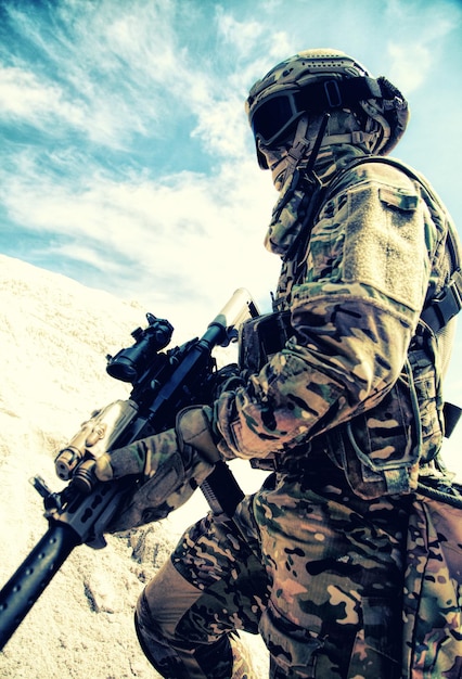 Airsoft, joueur de strikeball en uniforme de camouflage avec munitions tactiques militaires et répliques de carabine de service grimpant sur une dune de sable. Jeu de guerre moderne dans une zone sablonneuse ou dans le désert, imitation de conflit armé