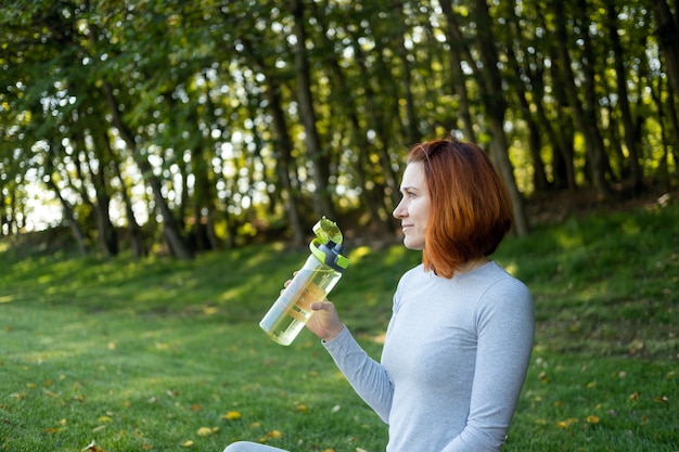 Ajuster la femme en vêtements de sport, boire de l'eau pendant l'entraînement, faire du yoga dans le parc par une journée ensoleillée d'automne.