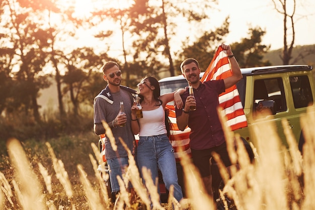 Photo avec de l'alcool dans les mains. les amis passent un bon week-end à l'extérieur près de leur voiture verte avec le drapeau américain.