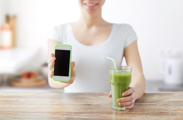 alimentation saine, régime alimentaire, désintoxication, technologie et concept humain - gros plan d'une femme avec du jus vert de smartphone assis à une table en bois sur fond de cuisine
