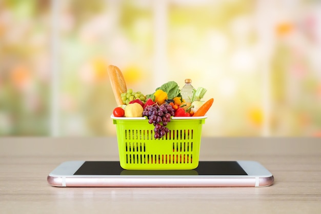 Aliments frais et légumes dans le panier vert sur smartphone mobile sur table en bois avec fenêtre et jardin abstrait flou fond épicerie concept en ligne