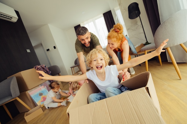 Photo aller de l'avant. homme heureux et positif poussant la boîte en carton avec son fils dedans tout en s'amusant