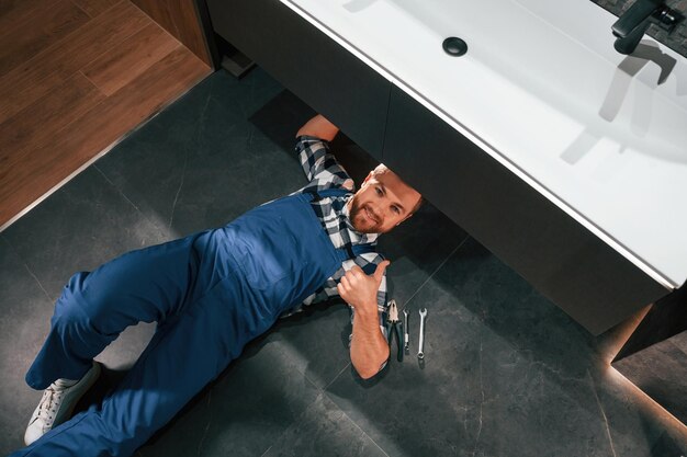 Photo allongé sur le sol fixant le siphon le plombier en uniforme bleu est au travail dans la salle de bain