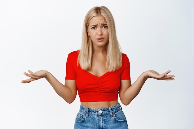 Alors quoi Agacée et confuse blonde fille arrogante haussant les épaules ne comprends pas quelque chose debout sur fond blanc