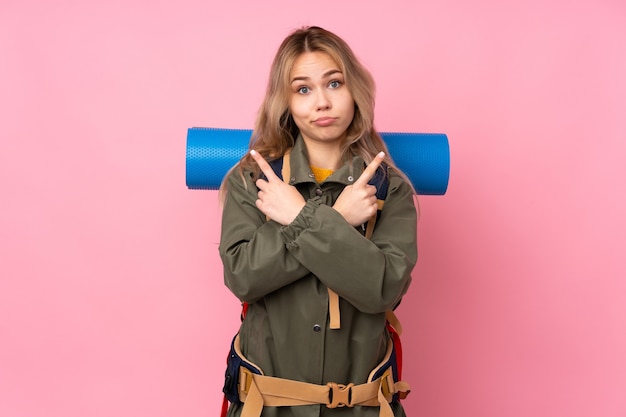Alpiniste russe adolescent fille avec un gros sac à dos isolé sur un mur rose pointant vers les latéraux ayant des doutes