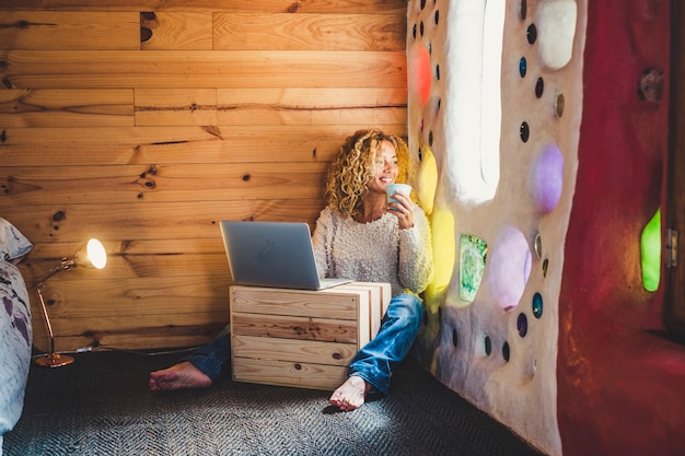 Alternative blonde d'âge moyen jolie femme s'asseoir dans la maison artistique et regarder par la fenêtre tout en faisant une pause avec du thé ou du café et utiliser un ordinateur portable pour les loisirs ou le travail