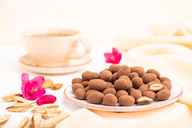 Amande dans des dragées au chocolat sur une plaque en céramique et une tasse de café sur fond de béton blanc et textile en lin orange. Vue latérale, gros plan, mise au point sélective.