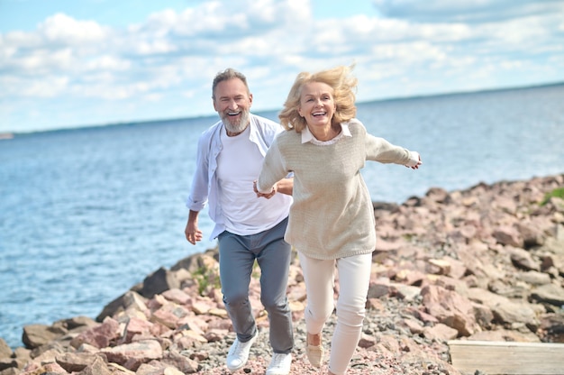 Ambiance délicieuse. Femme riante enthousiaste avec du vent dans ses cheveux tenant la main d'un homme barbu qui court le long du bord de mer aux beaux jours