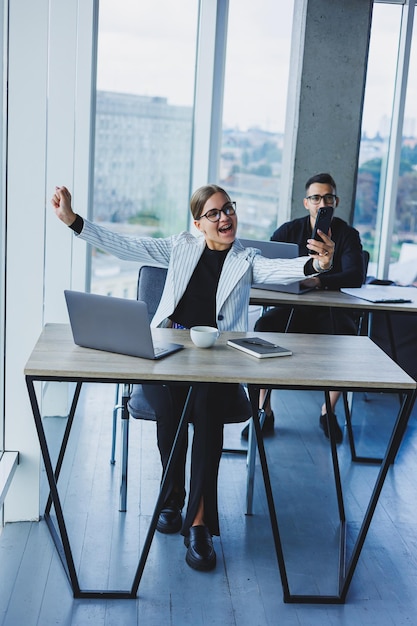 Ambiance de travail dans un bureau avec de grandes fenêtres Une femme gestionnaire est assise à un bureau à l'aide d'un ordinateur portable dans un bureau moderne dans le contexte d'un collègue