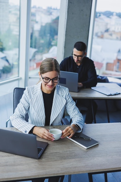 Ambiance de travail dans un bureau avec de grandes fenêtres Une femme gestionnaire est assise à un bureau à l'aide d'un ordinateur portable dans un bureau moderne dans le contexte d'un collègue