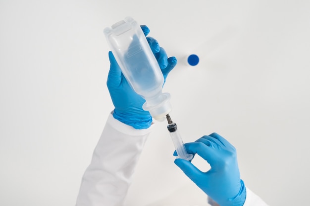 Un ambulancier sur une surface blanche dans des gants en latex bleu prend une solution pour séparer le médicament en poudre d'une grande bouteille transparente avec une seringue