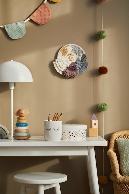 Aménagement intérieur d'une chambre d'enfant élégante avec étagère blanche, jouets en bois, poupées, accessoires pour enfants, lampe blanche, décoration confortable et drapeaux en coton suspendus sur le mur beige. Modèle.