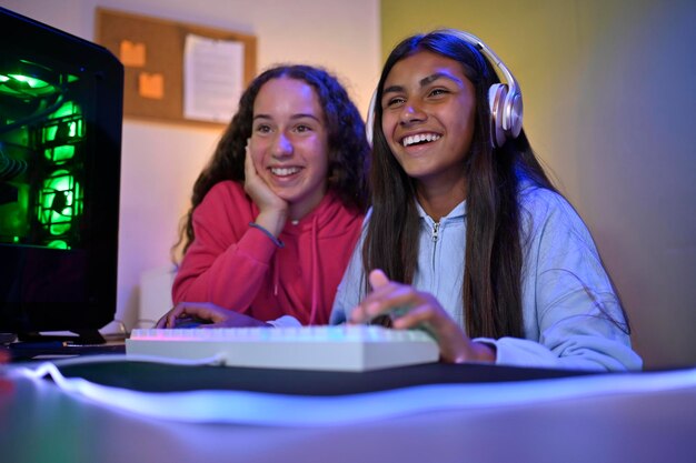 Des amies adolescentes multiraciales positives regardent l'écran du moniteur tout en étant assises à table avec un PC de jeu et en utilisant le clavier pour jouer au jeu dans la pièce