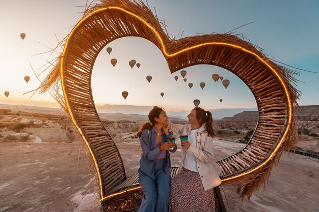 Photo amies assises sur un banc en forme de cœur décoré sur un point de vue et admirant la vue sur le vol de montgolfières en cappadoce