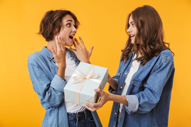 Amies de femmes isolées sur mur jaune tenant une boîte cadeau surprise.