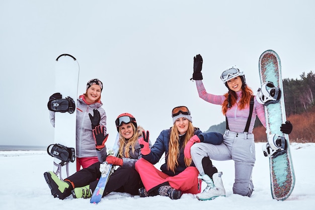 Amies gaies dans des vêtements d'hiver de sport avec des planches à neige et des skis se serrant les coudes et regardant une caméra tout en étant assis sur une plage enneigée