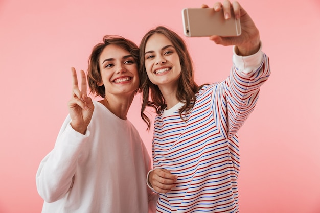 Les amies isolées sur le mur rose prennent un sefie par téléphone mobile.