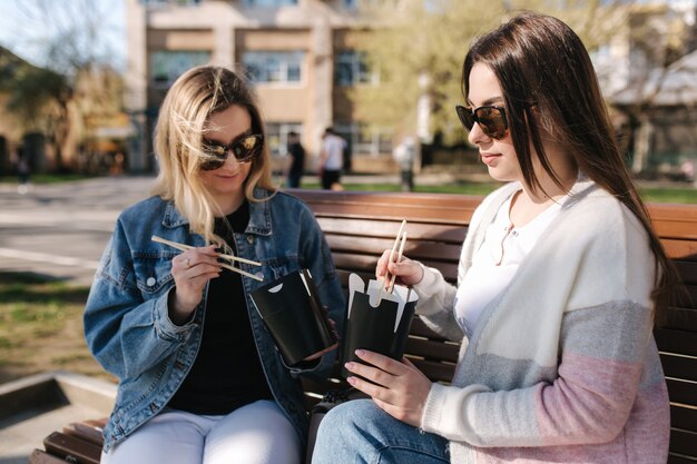 Des amies mangent des plats asiatiques à emporter assis sur un banc