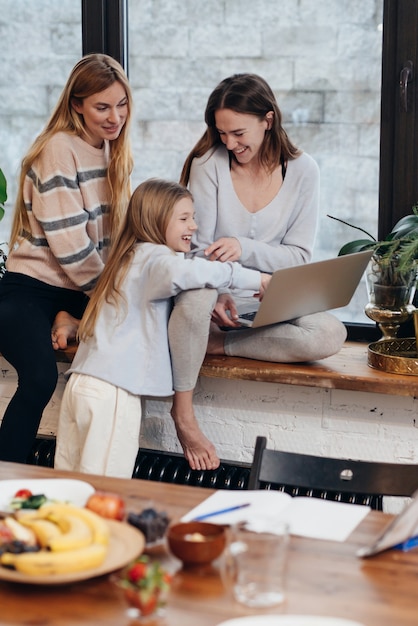 Des amies et une petite fille sont assises à la maison sur un rebord de fenêtre et regardent une série.