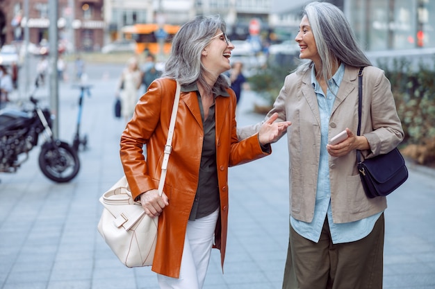 Des amies seniors positives parlent en marchant le long de la rue de la ville moderne