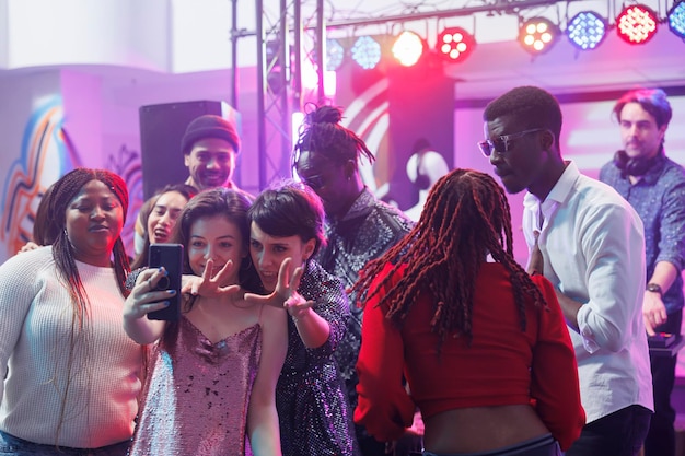 Des amies souriantes prenant un selfie sur leur smartphone tout en faisant la fête avec des amis dans une boîte de nuit. Des femmes joyeuses posant pour une photo sur leur téléphone portable tout en s'amusant à la discothèque dans un club.