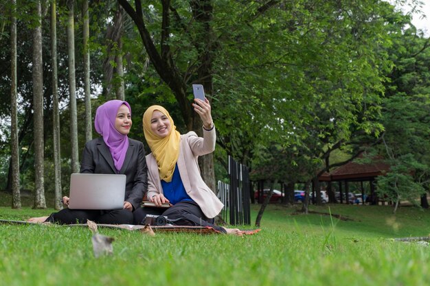 Photo des amies souriantes se font un selfie avec leur smartphone alors qu'elles sont assises sur un champ herbeux contre des arbres dans un parc.