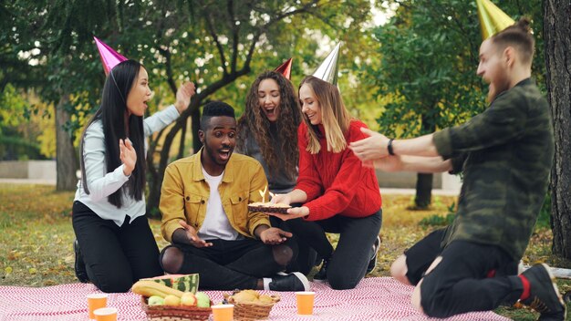 Des amis attentionnés apportent un gâteau à un homme afro-américain assis sur une couverture dans un parc en pique-nique les yeux fermés, il souffle des bougies et rit en profitant de la surprise