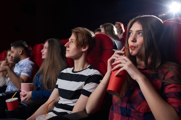 Amis et couples, regardant un film intéressant dans une grande salle de cinéma avec des chaises rouges, regardant sérieusement l'écran, pensant au film et buvant du cola. Concept de culture et de divertissement.