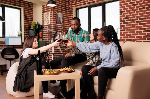 Amis de diverses cultures, afro-américains, asiatiques, caucasiens heureux célébration assis au salon de la maison. Les collègues portent un toast au vin, aux bâtonnets de pain et aux fromages.