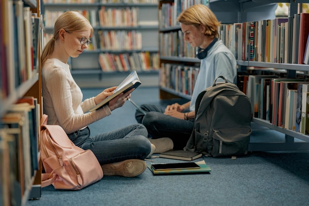 Des amis étudiants s'assoient sur le sol près des étagères de la bibliothèque et étudient en préparation à l'examen