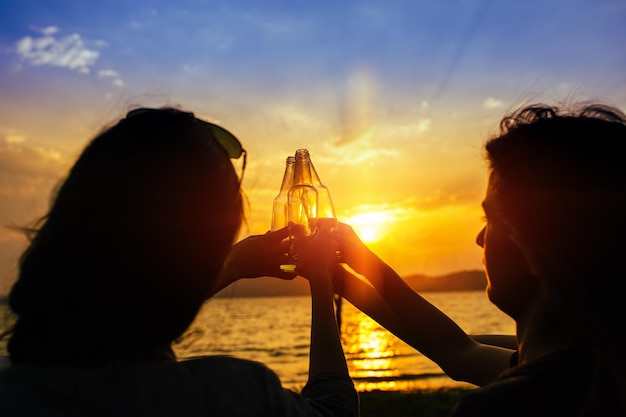 Photo des amis grillent des bouteilles de bière contre le lac au coucher du soleil.