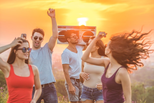 Les amis heureux avec une boîte de boom dansant sur le fond lumineux du soleil