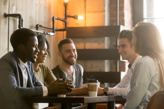 Des amis heureux de différentes races discutant et buvant du café, partageant une table de café, des jeunes du millénaire africains et caucasiens divers, souriants, appréciant une conversation agréable dans un café pendant la pause-café.