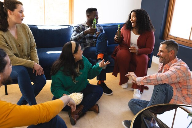 Photo des amis heureux et divers, hommes et femmes, qui parlent, boivent de la bière et mangent du pop-corn à la maison.