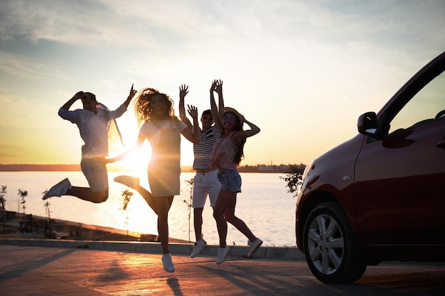 Amis heureux sautant près de la voiture à l'extérieur au coucher du soleil Voyage d'été