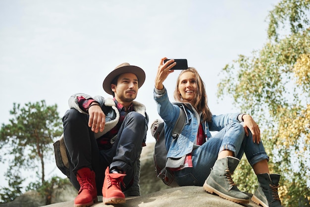 Photo des amis heureux se font un selfie assis sur un rocher dans la forêt.