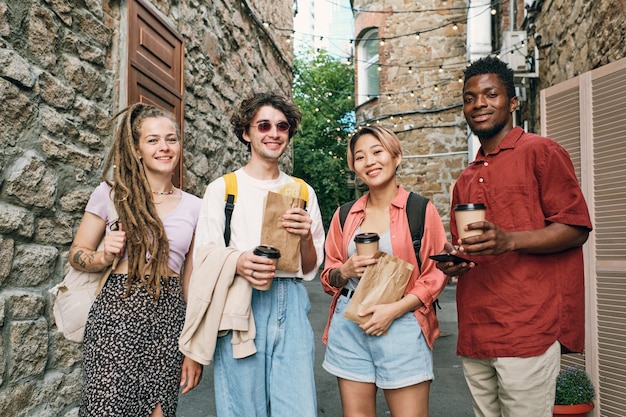 Amis interculturels heureux avec des boissons et des collations debout entre des bâtiments en pierre